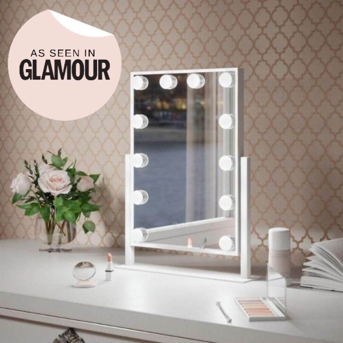 The Olsen Hollywood Mirror White 47x37cm by hollywood mirrors, featuring adjustable brightness and round LED bulbs, is displayed on a white desk with flowers, makeup, and perfume. A As Seen in GLAMOUR sticker appears on a beige patterned wall.