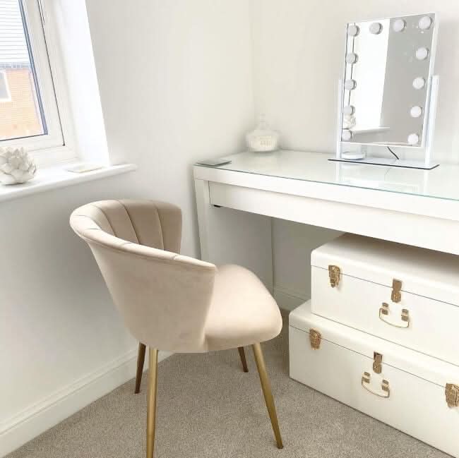 A white vanity with the hollywood mirrors Olsen Hollywood Mirror White 47x37cm, featuring adjustable brightness, sits by a window. A beige chair with gold legs is in front, and two white storage trunks are stacked beneath. The room has light walls and carpet.