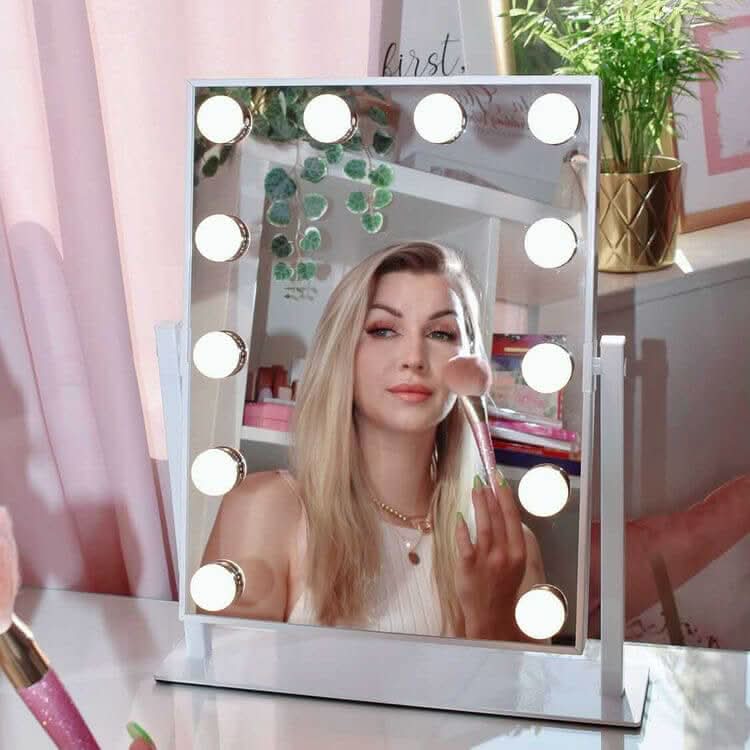 A woman with long blonde hair applies makeup using a brush, reflected in the hollywood mirrors Olsen Hollywood Mirror White 47x37cm with LED bulbs and adjustable brightness. Pink curtains and décor accent the background.