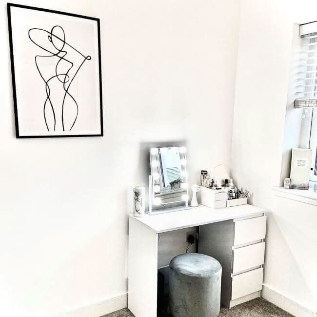 A small white vanity with drawers is paired with the hollywood mirrors Olsen Hollywood Mirror White 47x37cm, featuring adjustable LED bulbs, a gray round stool, makeup items, and a minimalist woman line art above, set near a window in a bright room.
