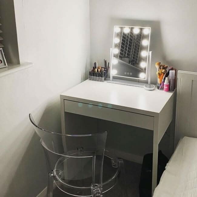 A compact white vanity table with the hollywood mirrors Olsen Hollywood Mirror White 47x37cm and LED bulbs, plus makeup brushes and cosmetics, sits by the bed and window with a clear acrylic chair.