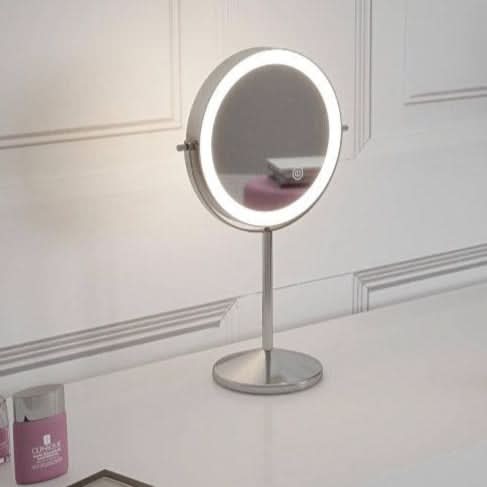 The Pre Order Mid June Illuminated Round Cosmetic Mirror (rechargeable, 20cm diameter) sits on a white vanity next to foundation, lipstick, eyeshadow palette, and pink perfume bottle against a white paneled wall.