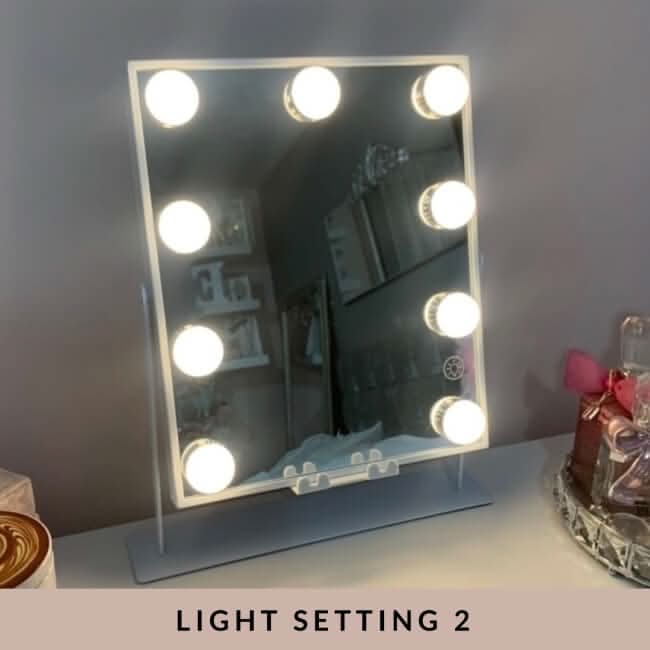 The Renée Bluetooth Hollywood Mirror 30x25cm with eight LED bulbs sits on a table, casting a bright glow. Its reflection shows shelves and decor. LIGHT SETTING 2 is displayed beneath the makeup mirror.
