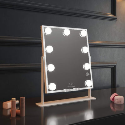 The Renée Bluetooth Hollywood Mirror 30x25cm, featuring eight LED bulbs, sits on a dark counter among makeup and skincare items, set against a paneled wall.