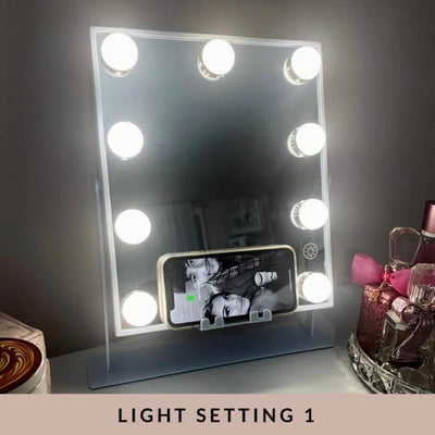 The Renée Bluetooth Hollywood Mirror 30x25cm with eight LED bulbs is illuminated on a desk. A smartphone sits in its base slot, showing a black-and-white image. Perfume bottles and beauty items are nearby. Text: LIGHT SETTING 1.