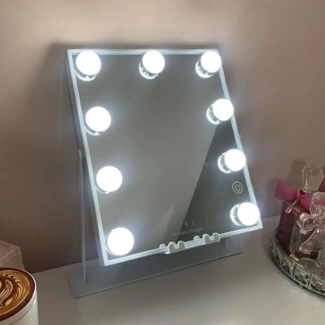 The Renée Bluetooth Hollywood Mirror 30x25cm, featuring eight round LED bulbs, sits slightly tilted backward on a table near perfume bottles and a decorative item.