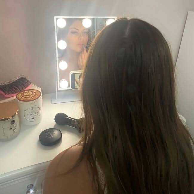 A woman with long brown hair applies lipstick at her vanity, looking into the Renée Bluetooth Hollywood Mirror 30x25cm with bright LED bulbs. Beauty products, a brush, and candles rest on the white tabletop. Her reflection is visible in the mirror.