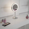 The Pre Order Mid June Illuminated Round Cosmetic Mirror (rechargeable, 20cm diameter) sits on a white vanity next to foundation, lipstick, eyeshadow palette, and pink perfume bottle against a white paneled wall.