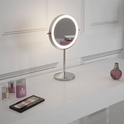 The Pre Order Mid June Illuminated Round Cosmetic Mirror (rechargeable, 20cm diameter) sits on a white vanity next to foundation, lipstick, eyeshadow palette, and pink perfume bottle against a white paneled wall.