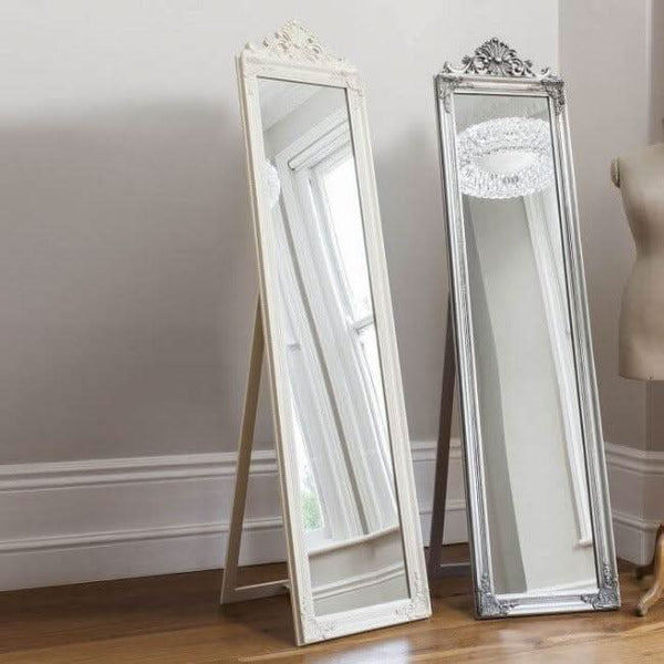 White wooden cheval floor mirror with decorative leaf finish, standing next to a silver mirror, in a well-lit room with wooden flooring.